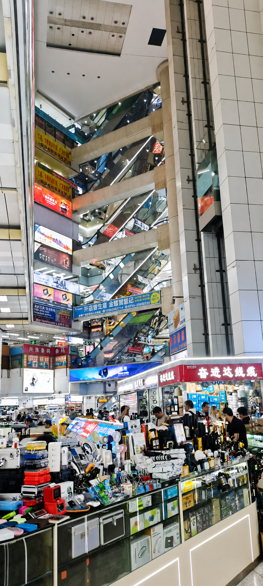 Bright professional interior photograph of Huaqiangbei electronics market counters, escalators, signage, and active supplier stalls.